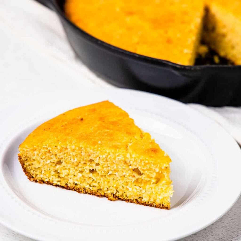 11 of the Best Side Dishes to Serve with Chili Golden cornbread slice on a white plate, cast-iron skillet behind