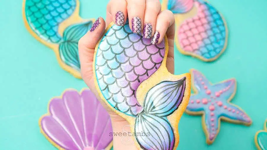 Hand holding pastel mermaid tail sugar cookie with scales, seashell and starfish iced cookies, matching nail art