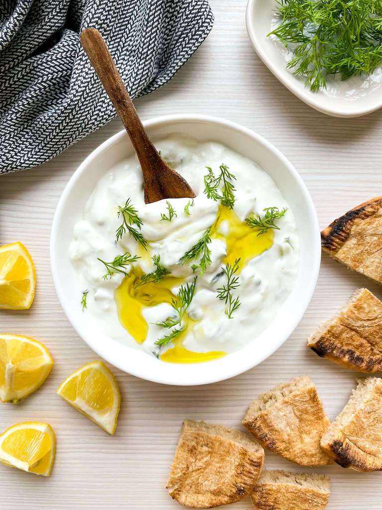 Creamy tzatziki sauce topped with fresh dill and olive oil with lemons, dill and pita bread for dipping.