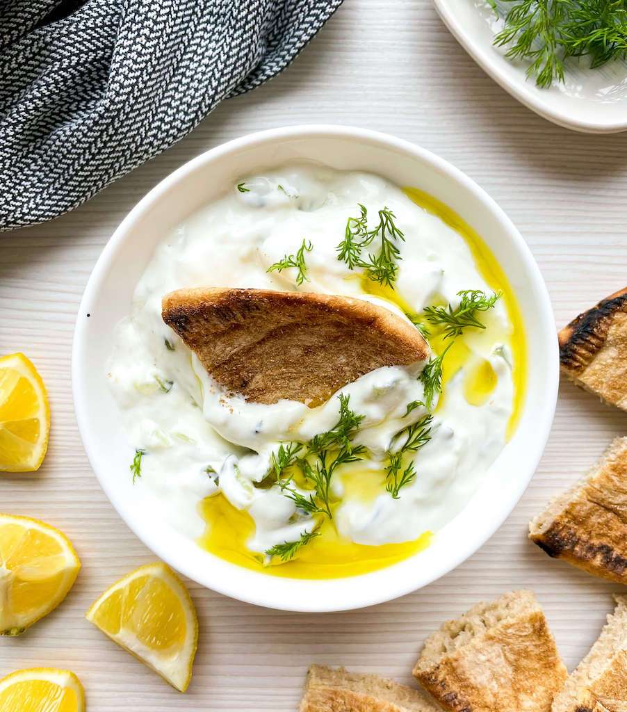 Creamy tzatziki sauce topped with fresh dill and olive oil with lemons, dill and pita bread for dipping.