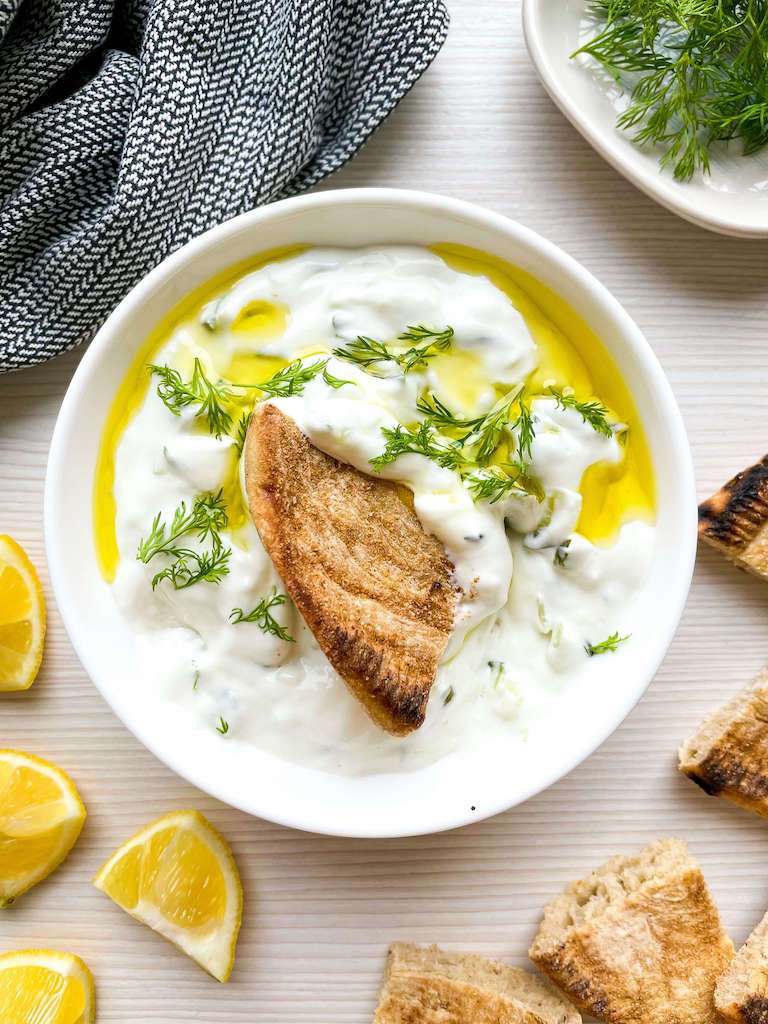 Tzatziki sauce topped with fresh dill and olive oil with lemons, dill and pita bread for dipping.