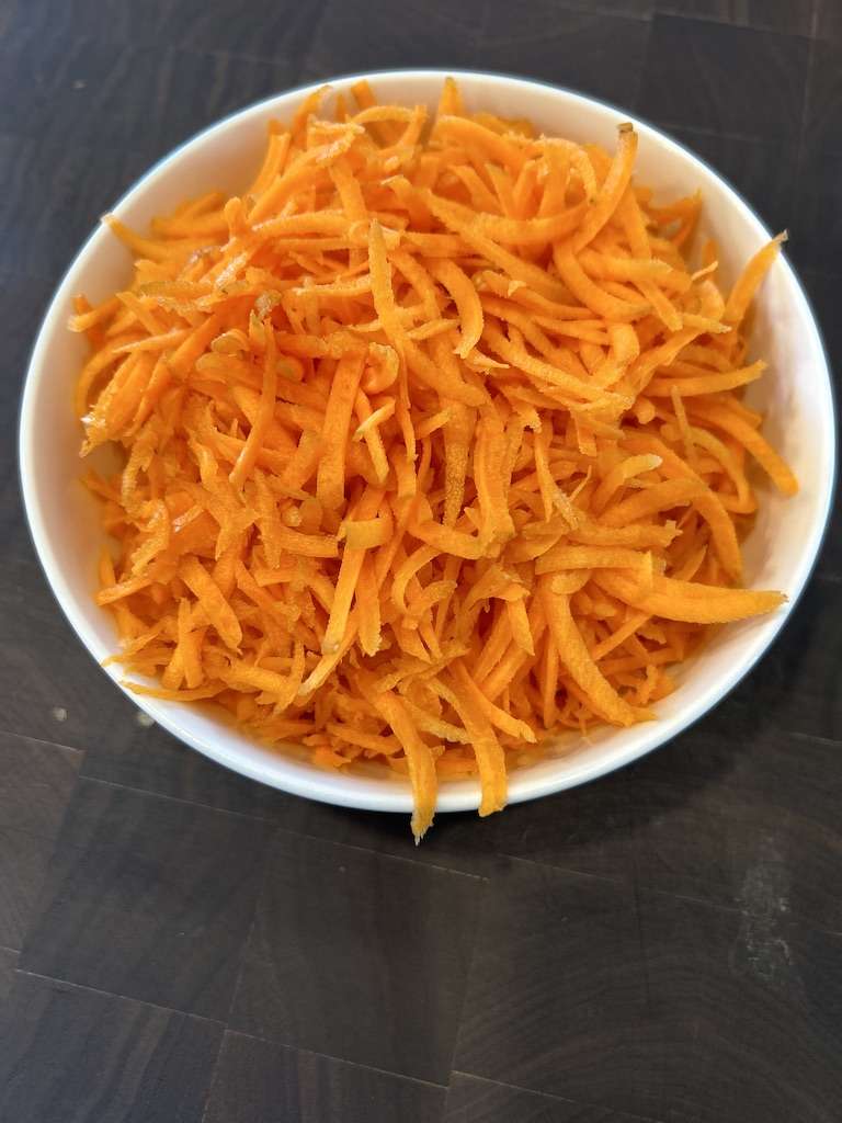 Grated Carrots in a white bowl on a wooden cutting board.