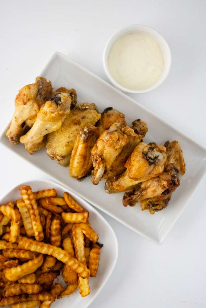 A white rectangular plate with lemon pepper wings in a row with french fries on the side in a white bowl.