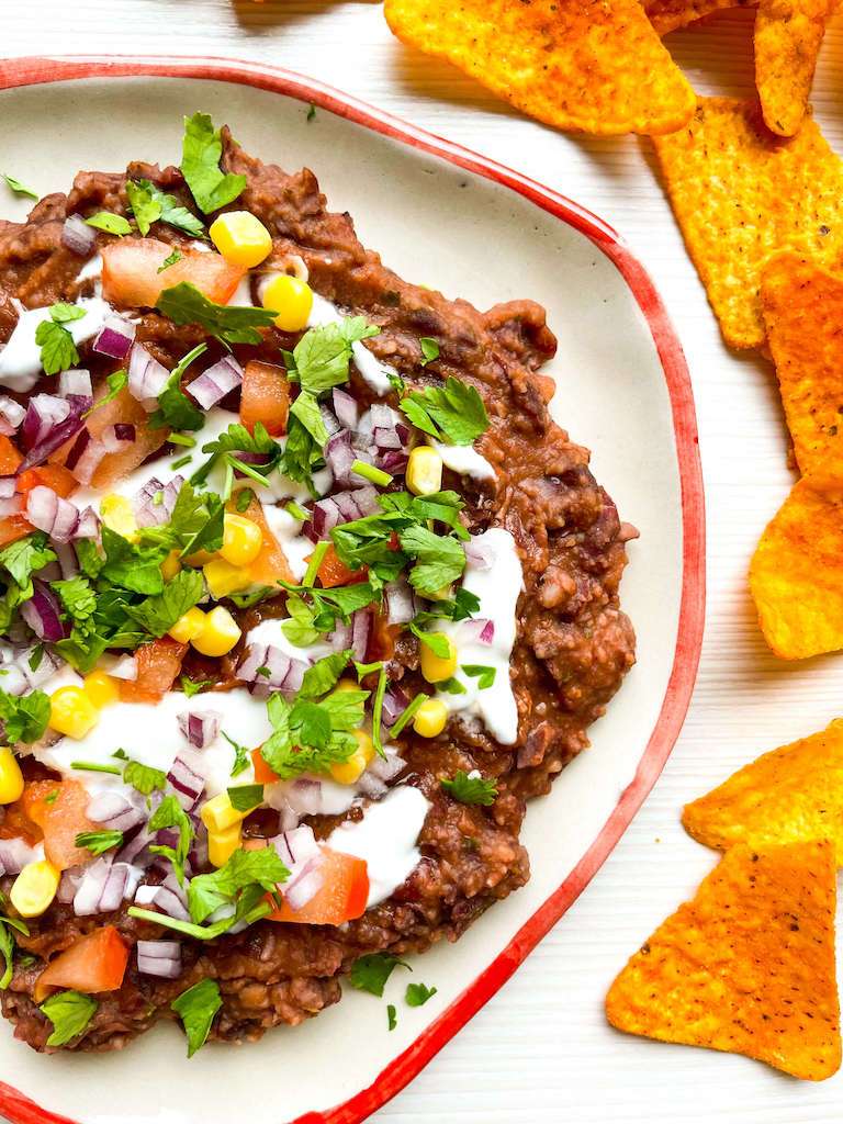 Loaded black bean dip garnished with fresh parsley, tomatoes, corn and red onion on a plate surrounded by chips.