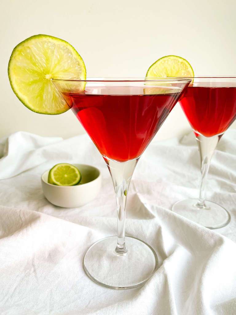Two pomegranate cosmopolitan drinks in martini glasses with lime slices for garnish.