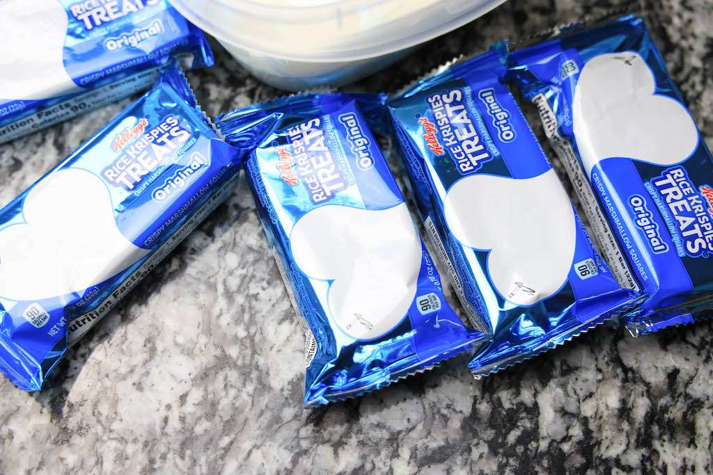 A few individually wrapped rice Krispie treats in their packages next to a bowl of melted white chocolate on a marble counter.