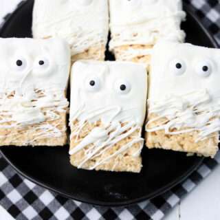 Five mummy rice krispie treats on a black plate with a black and white checkered napkin underneath the plate.