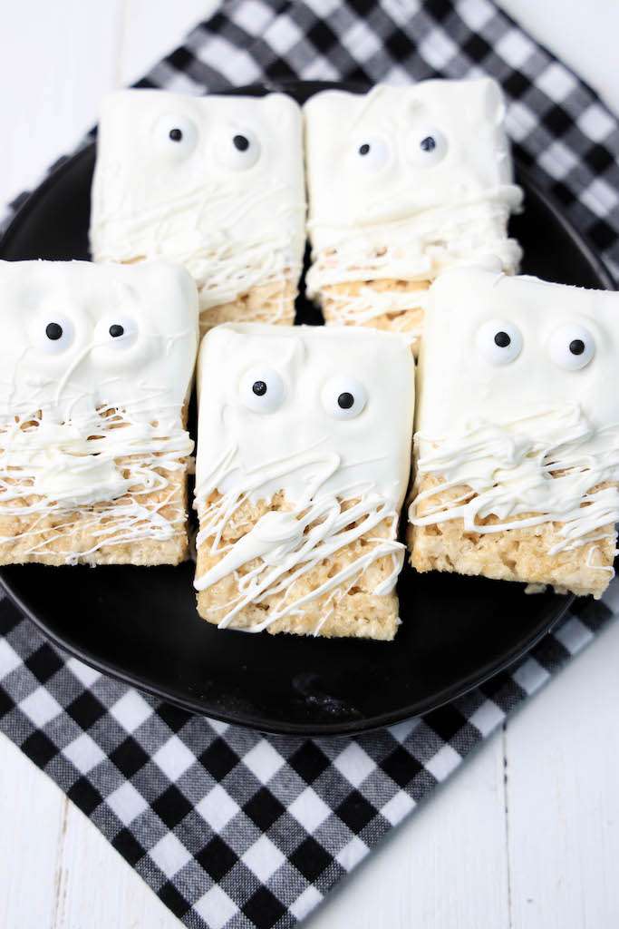 Five mummy rice krispie treats on a black plate with a black and white checkered napkin underneath the plate.