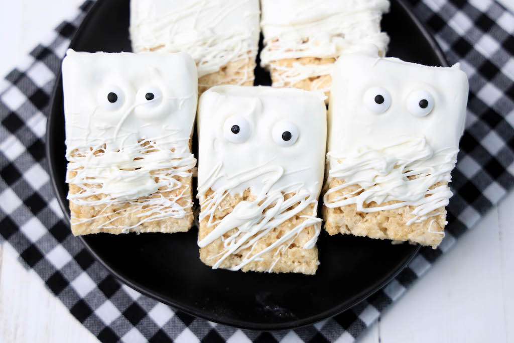 Three mummy rice krispie treats on a black plate with a black and white checkered napkin underneath the plate.