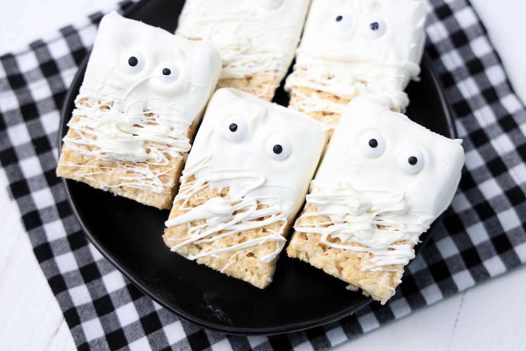 Four rice krispie mummies treats on a black plate with a black and white checkered napkin underneath the plate.