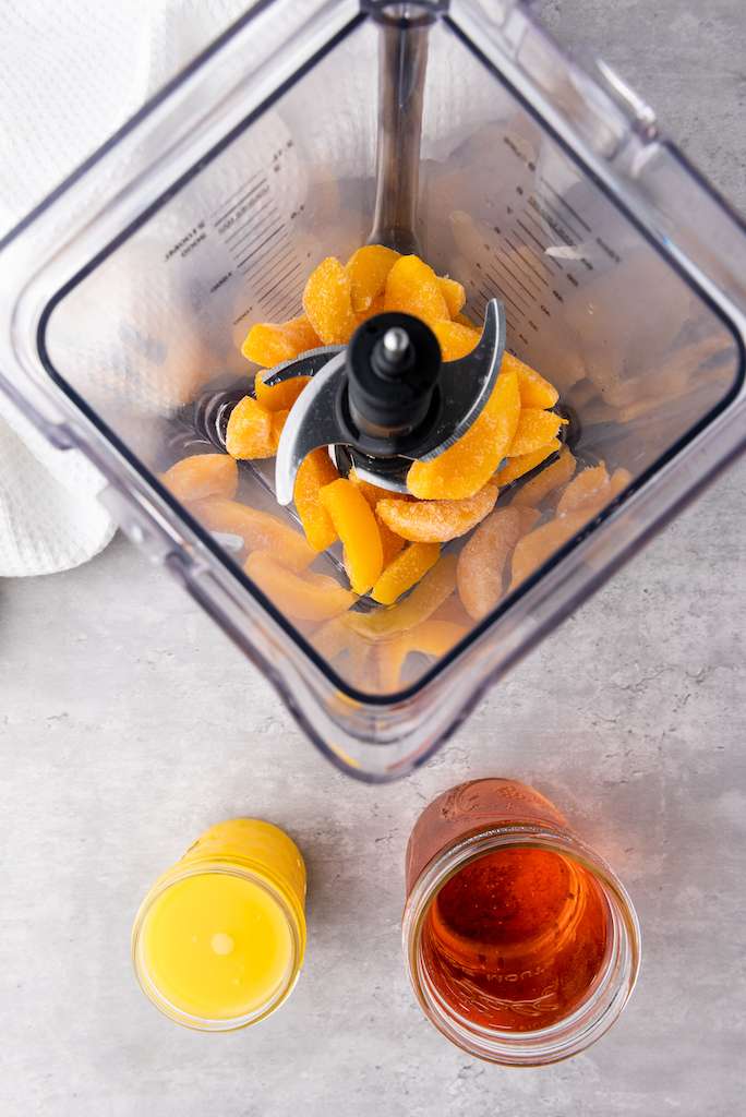 Frozen peach slices in a blender with orange juice and rose wine in glasses nearby to make wine pops.