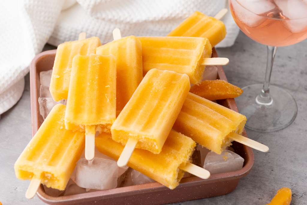 30+ Sweet Tooth Dessert Recipes Peach Rose Wine Popsicles on a copper cookie sheet with a glass of peach rose wine and a white towel in the background.