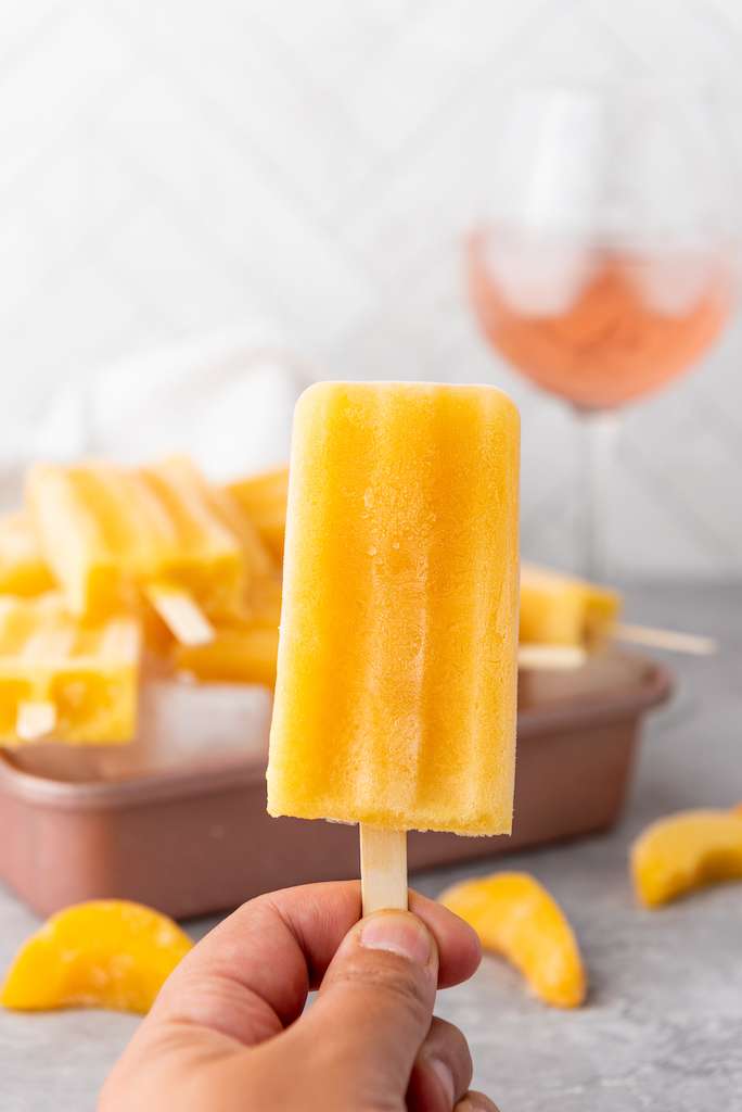 Peach Rose Wine Popsicles on a stick held up with Peach Rose Wine Popsicles on a copper cookie sheet with a glass of wine and a white towel in the background.