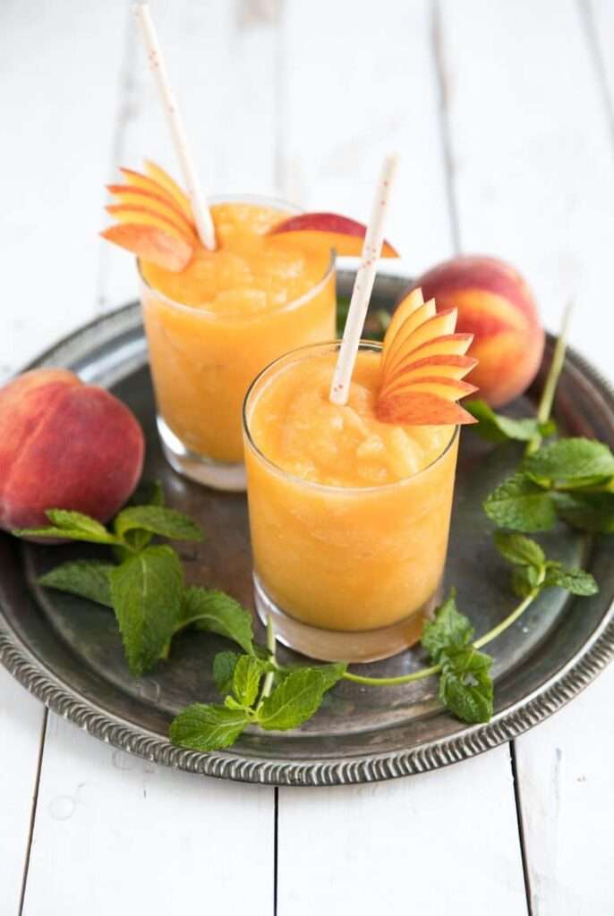 11 Refreshing Wine Slushie Recipes for Summer Peach smoothie slush in glasses with mint and peach slices on tray