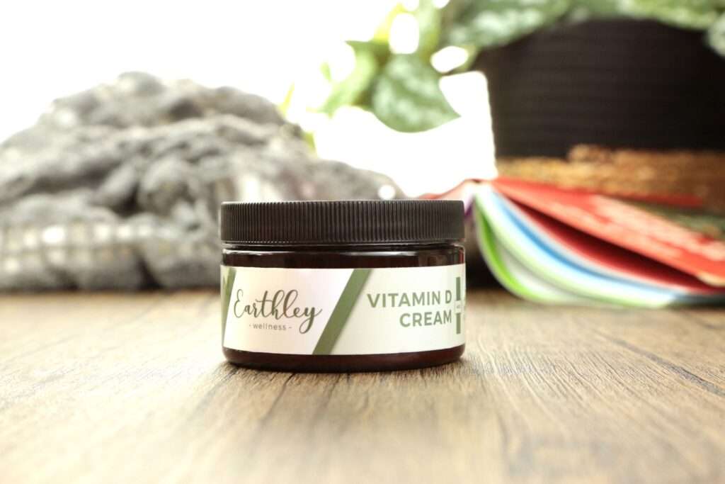 A jar of vitamin D cream on a wooden table with a blanket, plant and a book in the background.