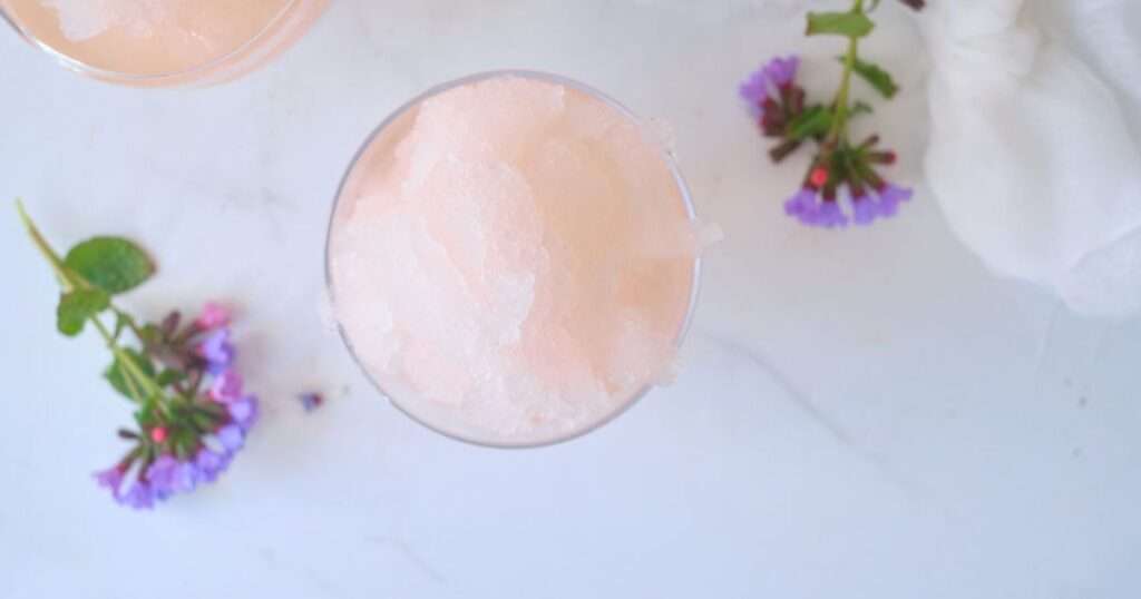 11 Refreshing Wine Slushie Recipes for Summer Pale pink granita slush in a glass on white marble, with purple flowers