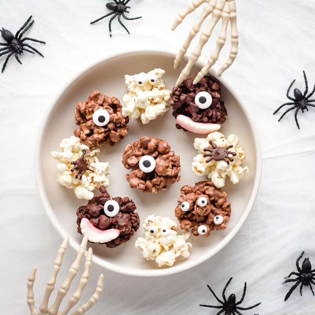 16 Easy & Spooky No Bake Halloween Treats Halloween popcorn monster treats with chocolate, candy eyes, spiders, and skeleton hands on plate.