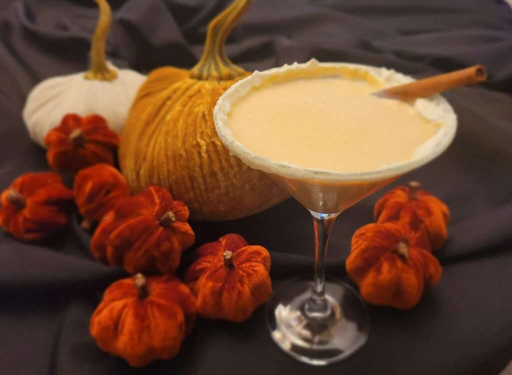 11 Delicious Pumpkin Cocktails to Try This Fall Creamy pumpkin pie martini with cinnamon stick and sugared rim, surrounded by pumpkins on dark background