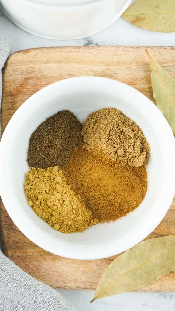 All 4 spices needed to make pumpkin pie spice in a small white bowl on a wooden cutting board.