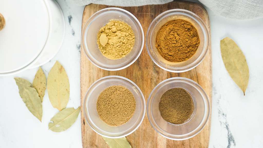 The 4 spices needed to make pumpkin pie spice in small glass bowls on a wooden cutting board.