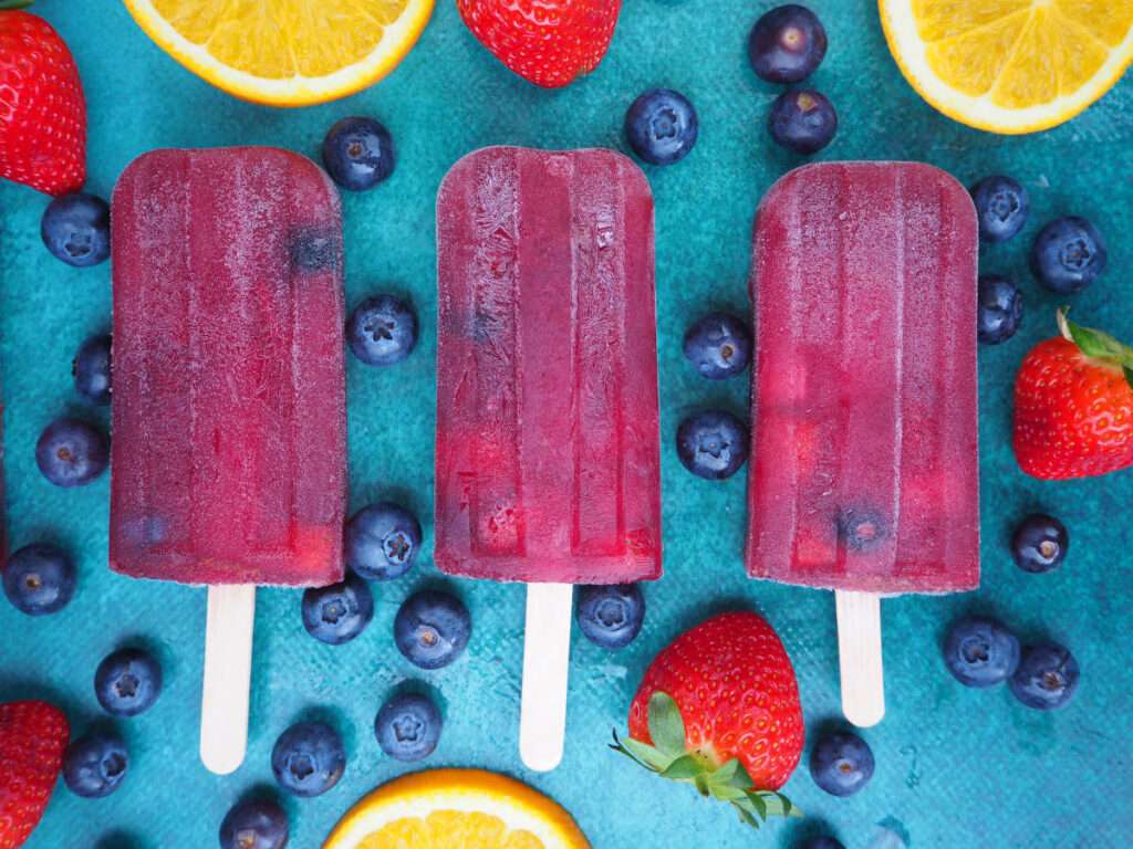 7 Delicious Wine Popsicle Recipes to Try This Summer Three homemade berry popsicles with blueberries, strawberries, and orange slices on teal background