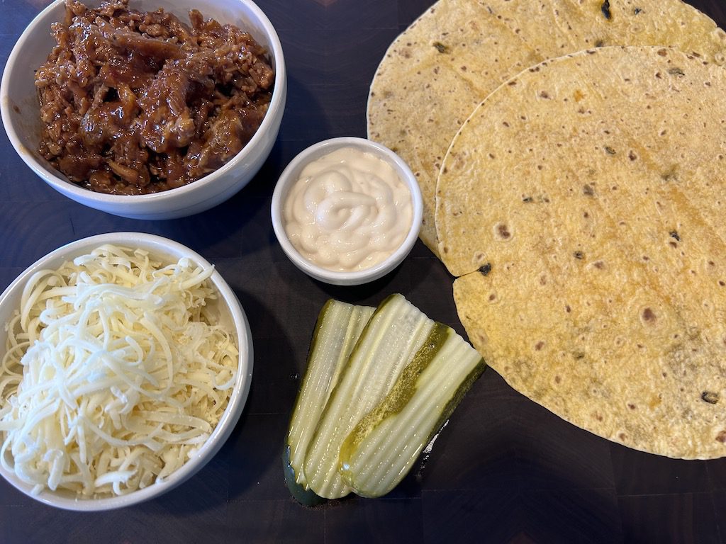 The ingredients for a Cuban quesadilla including barbecue shredded pork, jalenpeno cheddar tortillas, garlic aioli, shredded Swiss cheese, and Dill pickle slices on a wooden cutting board.