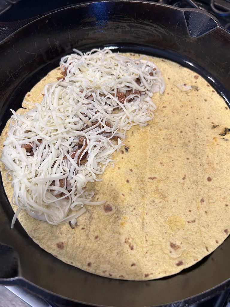 The Cuban quesadilla in the skillet with half covered with shredded cheddar.