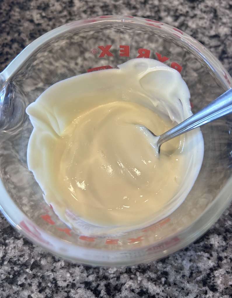 Melted white almond bark in a glass measuring cup with a silver spoon.