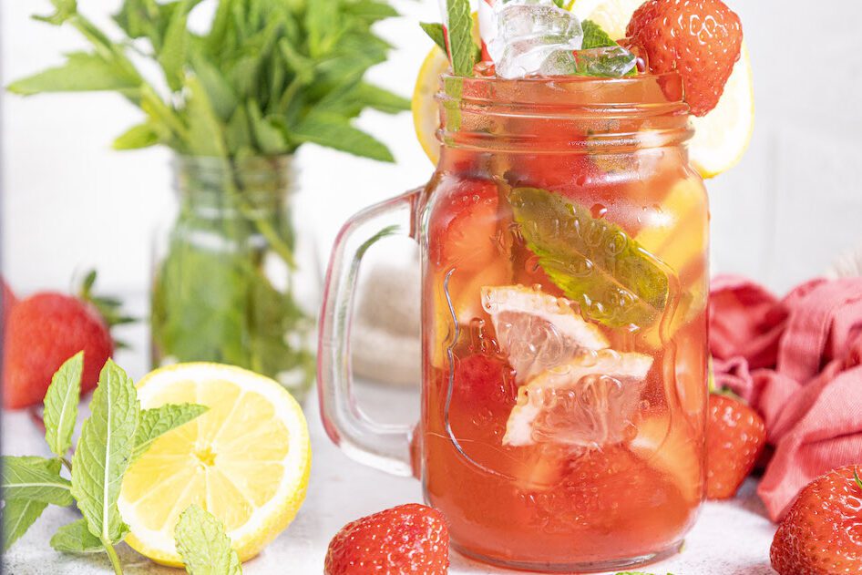 A mason jar glass filled with strawberry mojito mocktail garnished with fresh mint and strawberries with a class of mint leaves and fresh lemons around it.