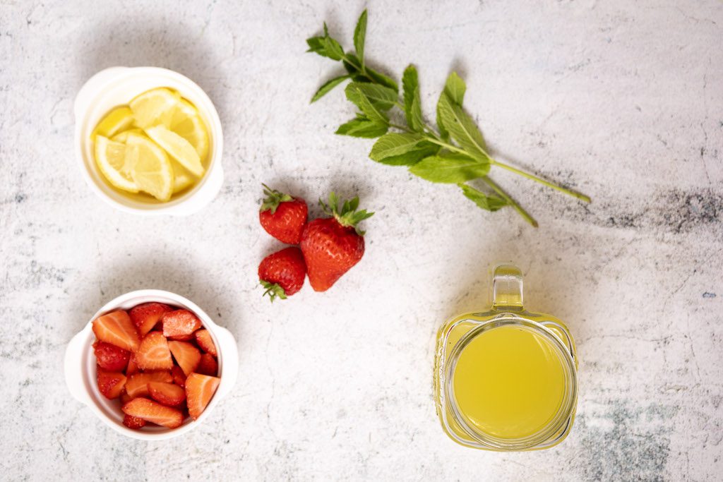 The ingredients for a nonalcoholic strawberry mojito including fresh sliced lemons, fresh strawberries, mint leaves, and sparkling water.