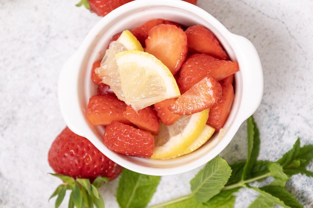 Fresh slices strawberries and lemon slices in a white bowl with strawberries and mint on the side.