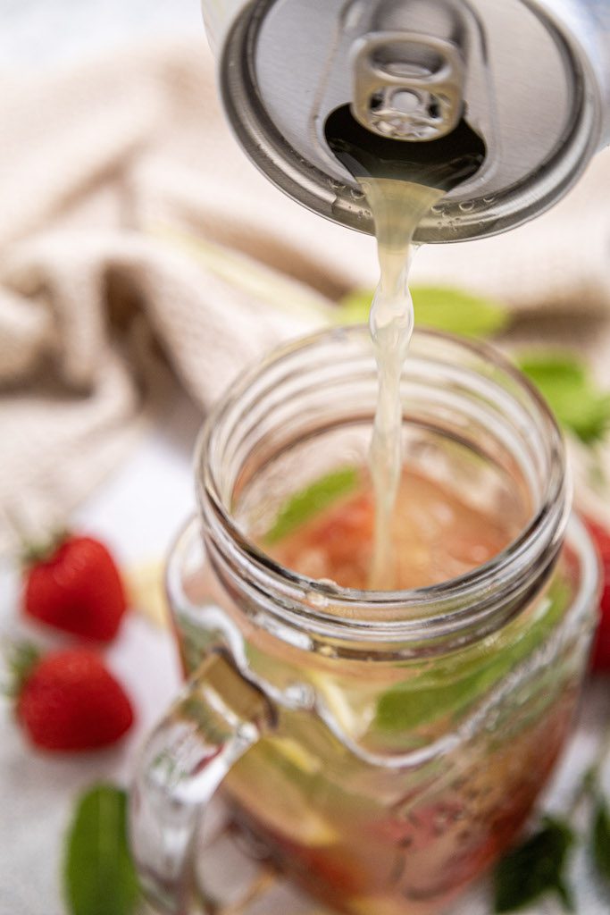 Pouring a can of sparkling water over a tall mason jar class of muddled strawberries, mint and lemon.