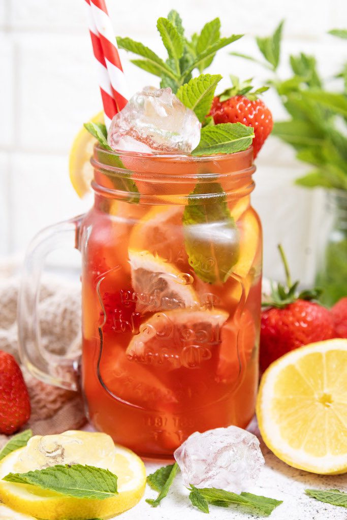 A mason jar glass filled with strawberry mojito mocktail garnished with fresh mint and strawberries with a class of mint leaves and fresh lemons around it.