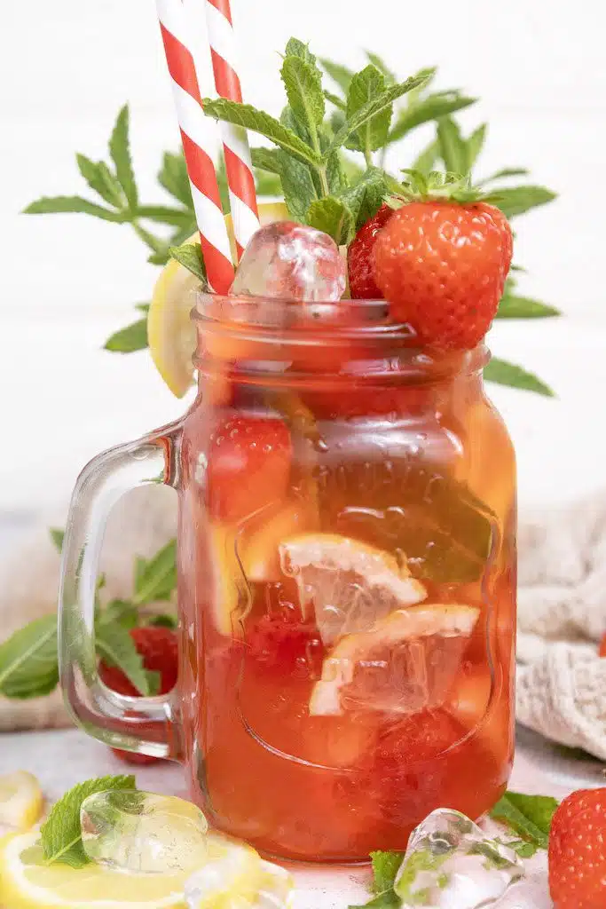 A mason jar glass filled with strawberry mojito mocktail garnished with fresh mint and strawberries with a class of mint leaves and fresh lemons around it.