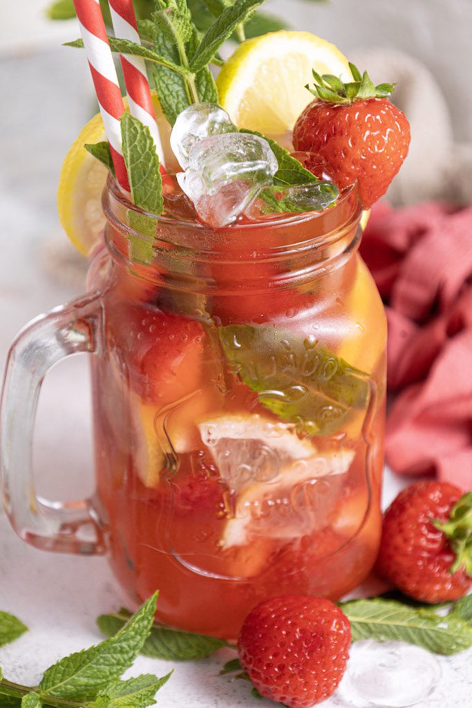 A mason jar glass filled with strawberry mojito mocktail garnished with fresh mint and strawberries with a class of mint leaves and fresh lemons around it.