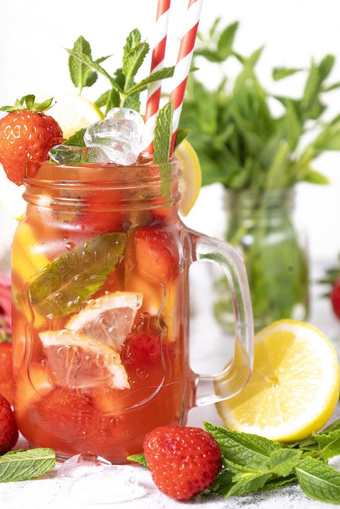 A mason jar glass filled with strawberry mojito mocktail garnished with fresh mint and strawberries with a class of mint leaves and fresh lemons around it.