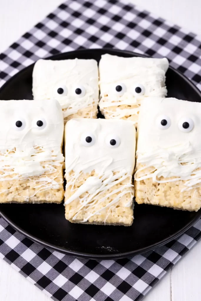 Halloween mummy Rice Krispies treats with candy eyes and white chocolate drizzle on a black plate