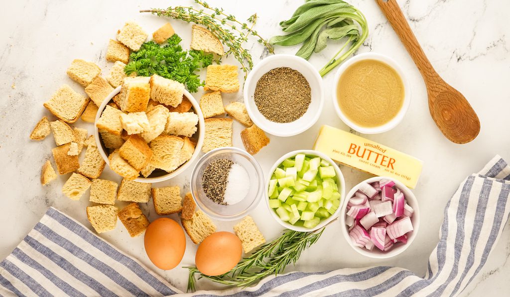 The ingredients for smoked stuffing including sourdough bread cubes, fresh herbs, seasonings, celery, red onion and chicken broth on a counter with a spoon.