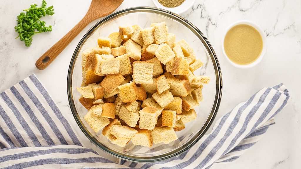 A large mixing bowl of sourdough bread cubes next to a striped towel, small white bowl of chicken broth and a wooden spoon.