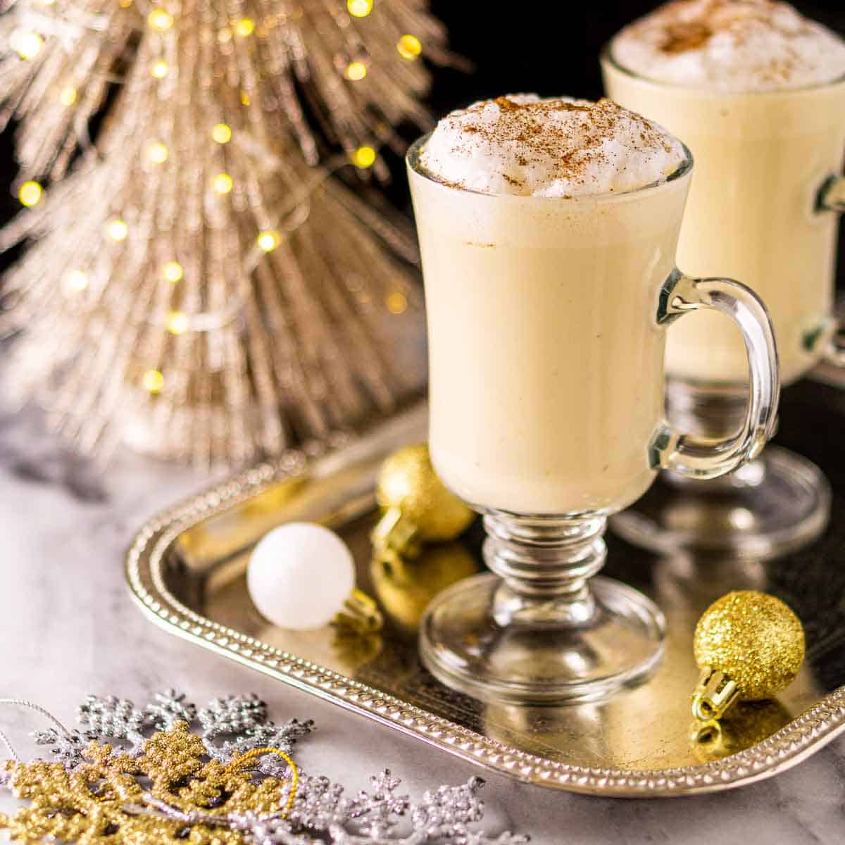 Baileys eggnog in a glass mug on a silver platter surrounded by gold and silver Christmas decorations.