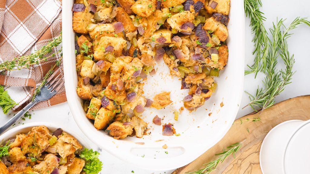 A serving of smoked stuffing next to the casserole dish of Thanksgiving smoked stuffing with a fork and fresh herbs.