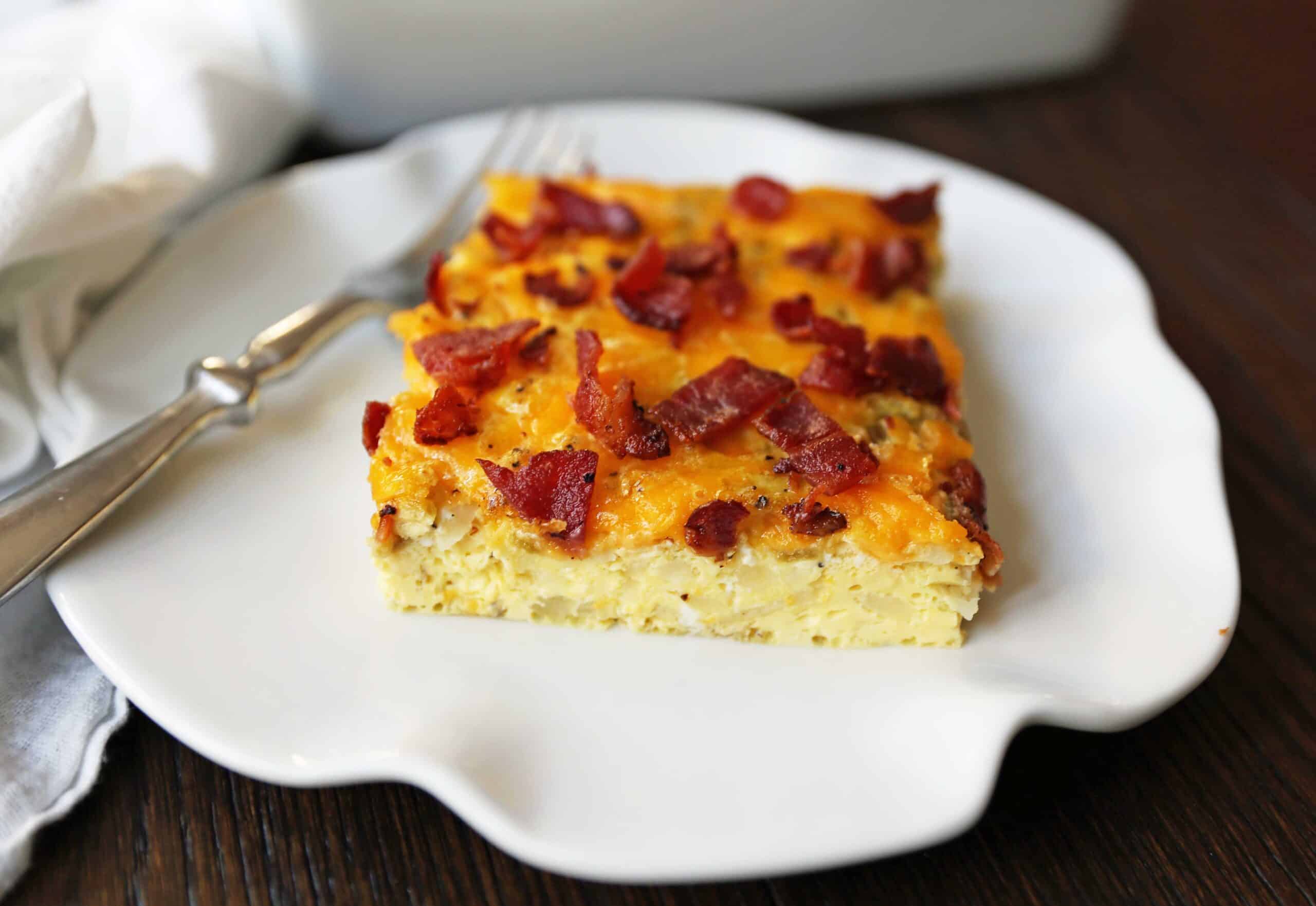 A slice of bacon, egg and cheese casserole o a white scalloped edged plate with a fork.