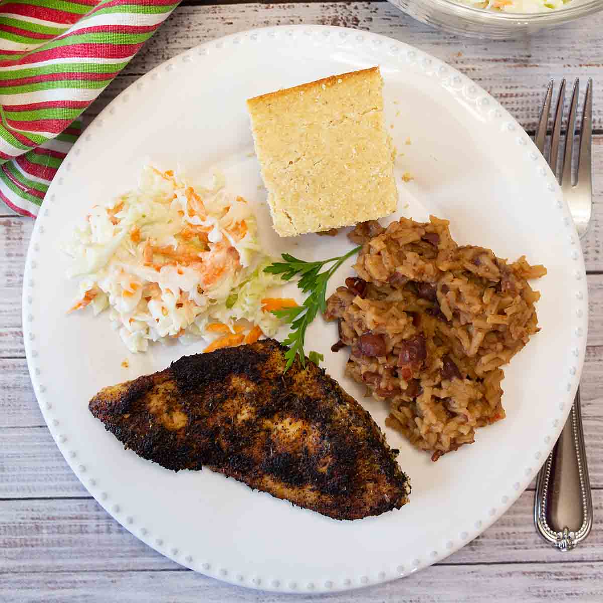 A white dinner plate featuring a blackened thinly sliced chicken breast, dirty rice, coleslaw, and corn bread.