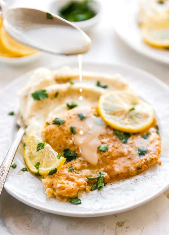 A white plate with chicken francese and fresh lemon slices with a spoon drizzling sauce over it.