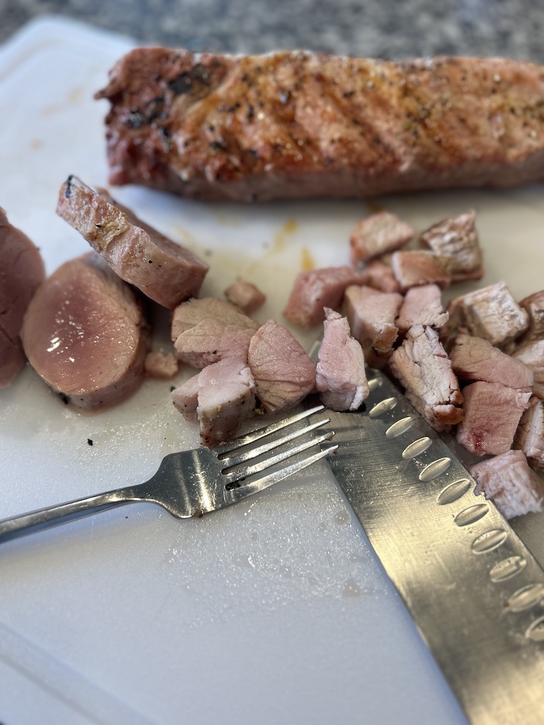 Cubes of pork tenderloin on a white cutting board with a knife and fork and an uncut smoked pork tenderloin in the background.