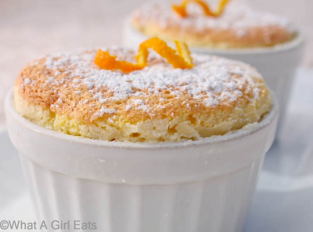 An egg souffle topped with a dusting of powdered sugar and a curl of orange peel in a white ramkin.