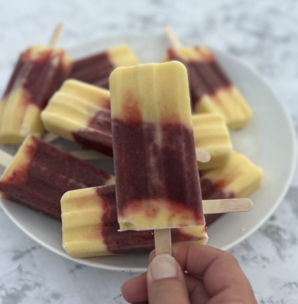 Someone holding a single mango cherry popsicle in front of a white plate piled with mango cherry popsicles.