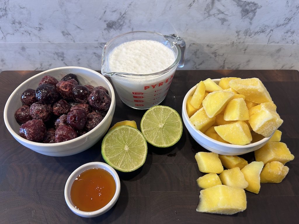 The ingredients for mango cherry popsicles including coconut milk, frozen cherries, frozen mangos and fresh limes.