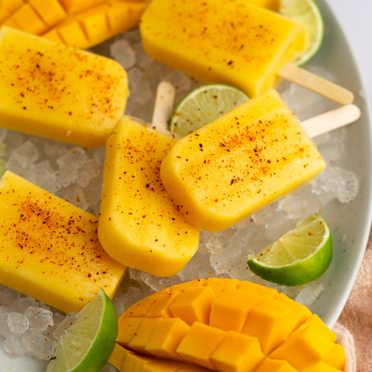 A few sugar free mango popsicles on ice in a white bowl with fresh lime wedges and a cubed mango.