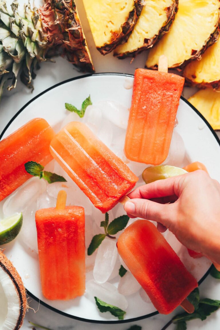 Five tropical fruit popsicles on a white black filled with ice with a sliced pineapple in the background.